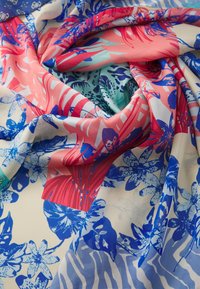Un tissu floral coloré présente de grands motifs tropicaux bleus, roses et verts sur un fond crème, avec une texture lisse et douce.