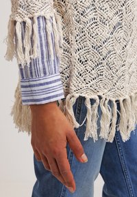 Gehaakte trui in room, met franjes; gelaagd over een blauw-witte gestreepte blouse met boorden. Textuur en lichtgewicht ontwerp.