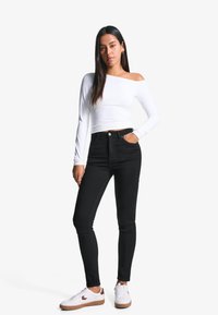 Svarta högmidjade jeans med smal passform, parat med en vit off-shoulder långärmad topp och vita sneakers med bruna detaljer.