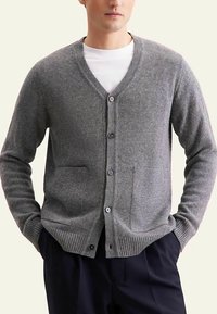 Cardigan gris en maille avec col en V, cinq boutons, deux poches avant, poignets et ourlet côtelés. Porté sur un t-shirt blanc.