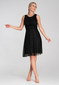 Schwarzes Kleid mit tailliertem Oberteil und ausgestellt Rock, überlagert mit transparenter Tüll. Verfügt über einen runden Ausschnitt und eine glatte Stoffstruktur.