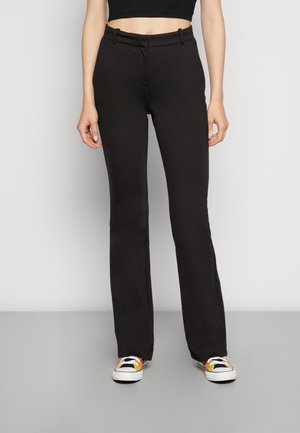 Mujer con pantalones negros de tiro alto y pierna recta y zapatillas coloridas con cordones, de pie frente a un fondo gris liso.