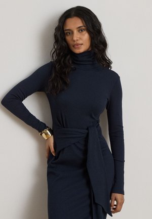 Donna con lunghi capelli ricci che indossa un vestito dolcevita blu navy con dettaglio annodato in vita e braccialetto dorato, posando davanti a uno sfondo semplice.