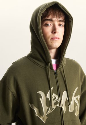 Sweat à capuche zippé vert olive avec un large motif crème sur le devant, doté d'une capuche à cordon et d'une texture de tissu douce.