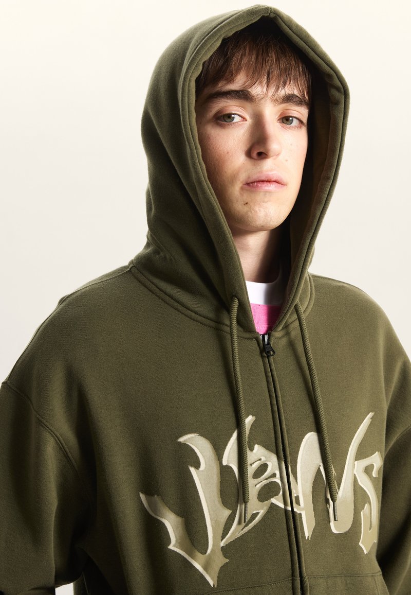 Sweat à capuche zippé vert olive avec un large motif crème sur le devant, doté d'une capuche à cordon et d'une texture de tissu douce.