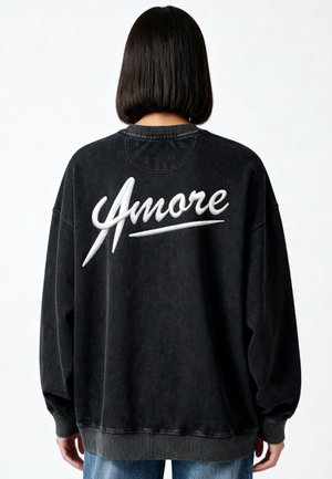 Personne aux cheveux foncés aux épaules porte un sweat-shirt noir oversize avec une grande inscription blanche "Amore" dans le dos, associé à un jean bleu.
