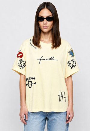 Donna che indossa una t-shirt giallo pallido con stampe grafiche nere e rosse e testo, abbinata a jeans azzurri e occhiali da sole neri, su sfondo bianco.