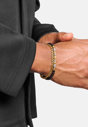 Zwei Armbänder an einem Handgelenk: eine Goldkette und eines mit schwarzen, goldenen und silbernen Perlen, getragen zu einem schwarzen Langarmshirt.