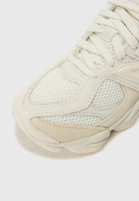 Lysfarvet sneaker med mesh-paneler, ruskindsdetaljer og teksturerede såler. Snørerne er vævet, og designet har buede linjer gennem hele skoen.