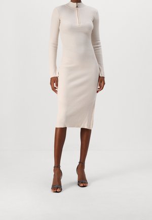 Kobieta w beżowej dzianinowej sukience midi w prążki z długimi rękawami, dekoltem na zamek błyskawiczny z przodu oraz czarnymi sandałami na wysokim obcasie z paskiem wokół kostki.