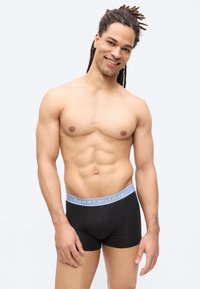 Χαμογελαστός νέος με dreadlocks στέκεται γυμνόστηθος φορώντας μαύρα μποξεράκια Tommy Hilfiger, σε λευκό ουδέτερο φόντο.