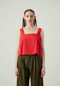 Jovem com cabelo trançado, vestindo uma blusa vermelha de decote quadrado sem mangas e calças largas de cintura alta em verde-oliva, contra um fundo simples.
