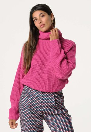 Overmaatse roze gebreide trui met een hoge col en geribbelde textuur, gecombineerd met een patroonbroek met geometrische vormen in blauw, rood en wit.