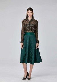 Blouse transparente marron à pois verts, associée à une jupe verte foncée longueur genoux avec une texture florale, portée avec des talons noirs.