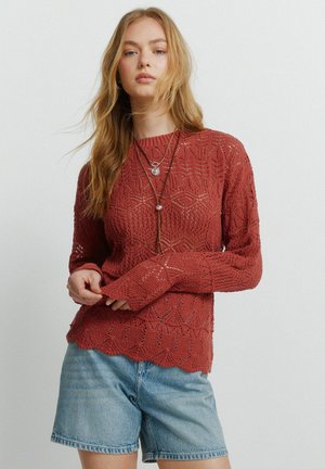 Jonge vrouw met lang haar, gekleed in een roestkleurige kanten gebreide trui en lichtblauwe spijkershorts, poserend tegen een effen achtergrond.