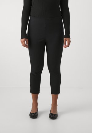 Persoon die zwarte cropped slim-fit broek, zwarte geribde top met lange mouwen en zwarte balletschoenen draagt, staand op een witte vloer.