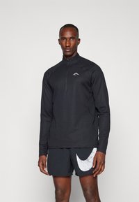Svart langermet jakke med glidelås i teksturert stoff og en liten hvit logo. Kombinert med svarte shorts som har en stor hvit Nike-swoosh.