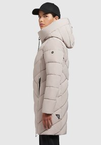 Gevoerde lange jas in lichtbeige met een capuchon, diagonale quiltpatroon, ritszakken en een logopatch aan de onderzijde.