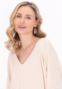 Top tricotat bej cu decolteu în V și mâneci lungi; prezintă o țesătură texturată. Modelul poartă cercei în formă de lacrimă cu un model pentru un detaliu suplimentar.