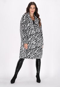 Cappotto in pelliccia sintetica con strisce zebra bianche e nere, dotato di un ampio colletto, chiusura con bottoni e vestibilità ampia. Abbinato a leggings lucidi neri.