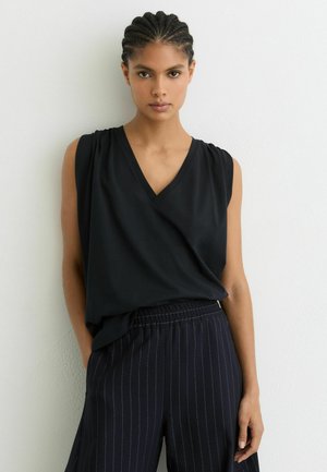 Młoda kobieta z warkoczami, ubrana w czarną, bezrękawną bluzkę z dekoltem w serek oraz czarne spodnie w cienkie paski, stojąca na tle białej ściany.