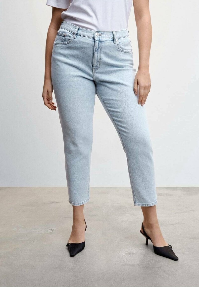 Femme portant un jean taille haute bleu clair, un t-shirt blanc et des talons pointus noirs, debout sur un sol en béton devant un mur uni.