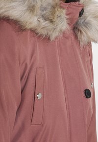 Parka rose poussière avec capuche en fausse fourrure, présentant une texture lisse, deux poches avant et des accents de boutons argentés et noirs.