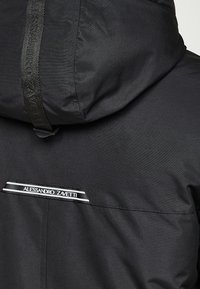 Černá kapucová bunda vyrobená z hladké tkaniny. Obsahuje značkovou nášivku a vodorovný pruh na zadní straně. Jednoduchý design, bez vzorů.