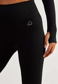 Black athletic leggings with textured fabric, high waist design, and a white heart logo on the thigh. Features thumbholes on the sleeves.