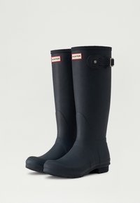 Botas de lluvia de goma negras con puntera redondeada, presentan una correa con hebilla en el lado y etiquetas "HUNTER" en rojo. Superficie lisa y texturizada.