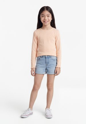 Young girl standing, wearing a peach long-sleeve shirt, light blue denim shorts, and white sneakers, smiling at the camera.
