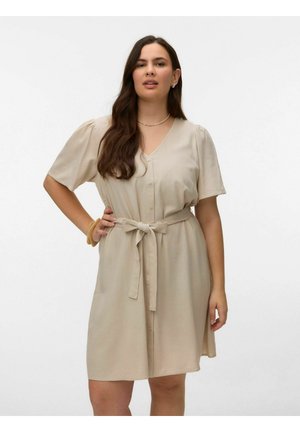 Donna con capelli lunghi e scuri che indossa un vestito beige con bottoni al ginocchio, maniche corte e cintura annodata in vita su uno sfondo bianco.