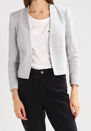 Femme portant un blazer gris clair sur un t-shirt blanc et un pantalon noir taille haute, debout devant un fond uni.