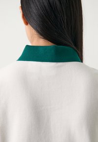 Top branco com gola canelada verde, feito de um tecido texturizado. Enfoque na vista traseira que revela o detalhe da gola.