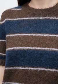 Pleteni džemper s vodoravnim prugama u smeđoj, tamnoplavoj i bijeloj boji. Mekana, pahuljasta tekstura s okruglim izrezom i kratkim rukavima.