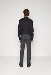 Chemise noire  à boutons associée à un pantalon gris à carreaux et à des bottes noires. La tenue présente une coupe slim avec un tissu lisse et des détails subtils.