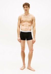 Czarne bawełniane szorty bokserskie z białym elastycznym paskiem w talii, na którym znajduje się logo "TOMMY HILFIGER" w czarnych literach. Model stoi na bosaka.