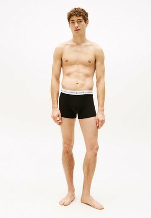 Zwarte katoenen boxershorts met een witte elastische tailleband waarop het logo "TOMMY HILFIGER" in zwarte letters staat. Model staat op blote voeten.
