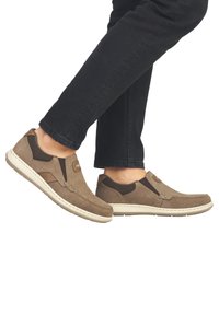 Bruine suède instappers met elastische zijkanten en beige rubberen zolen, met stiksels en een klein logo op de bovenkant.