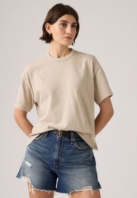 Γυναίκα που φοράει μια μπεζ φαρδιά κοντομάνικη μπλούζα και σκισμένα μπλε τζιν σορτς με μαύρη ζώνη, όρθια με τα χέρια πίσω από την πλάτη της.