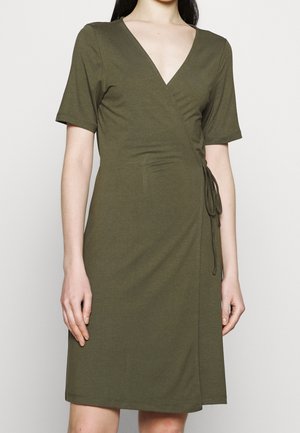Femme portant une robe portefeuille olive verte mi-longue avec manches courtes et détail de lien sur le côté, debout devant un fond uni.