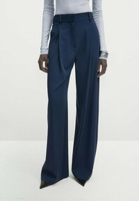 Pantalon large marine en tissu lisse, avec plis devant, taille ajustée et coupe épurée, porté avec des chaussures noires pointues.