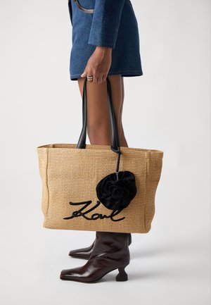 Sac au tissage beige avec des poignées en cuir noir, orné d'une rose en tissu noir et portant "Karl" en lettres cursives sur le devant.