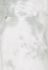 Valkoinen denim-kangas, jossa on hillitty harmaa tie-dye-kuvio. Sisältää takataskun, jossa on valkoinen ommel ja pieni metallinen nappi.