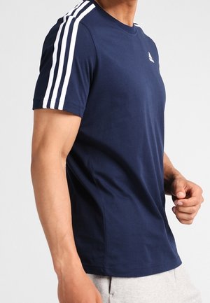 Mężczyzna w granatowej koszulce Adidas z białym wzorem trzech pasków na rękawach oraz małym logo na piersi, do tego jasnoszare spodnie.