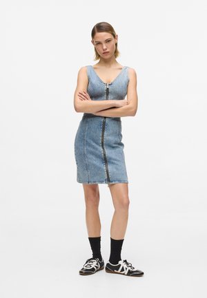 Rochie din denim cu croială mulată, decolteu în V, închidere frontală cu fermoar și lungime până la mijlocul coapsei. Purtați cu șosete și pantofi negri.