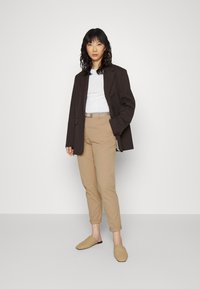 Brun blazer, hvit t-skjorte, beige skreddersydde bukser og vevde sandaler. Glatt stoff med oppbrettede ermer og subtil beltedetalj. Nøytral fargepalett.