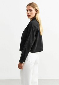 Cazadora negra corta de tela suave con mangas largas y detalle de botón. Combinada con pantalones blancos de pierna ancha, que muestran un ajuste relajado.