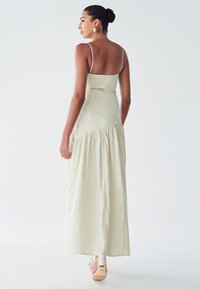 Robe longue beige clair avec des fines bretelles et un corsage ajusté, présentant une partie inférieure fluide et à niveaux, ainsi qu'une fermeture éclair dissimulée au dos.