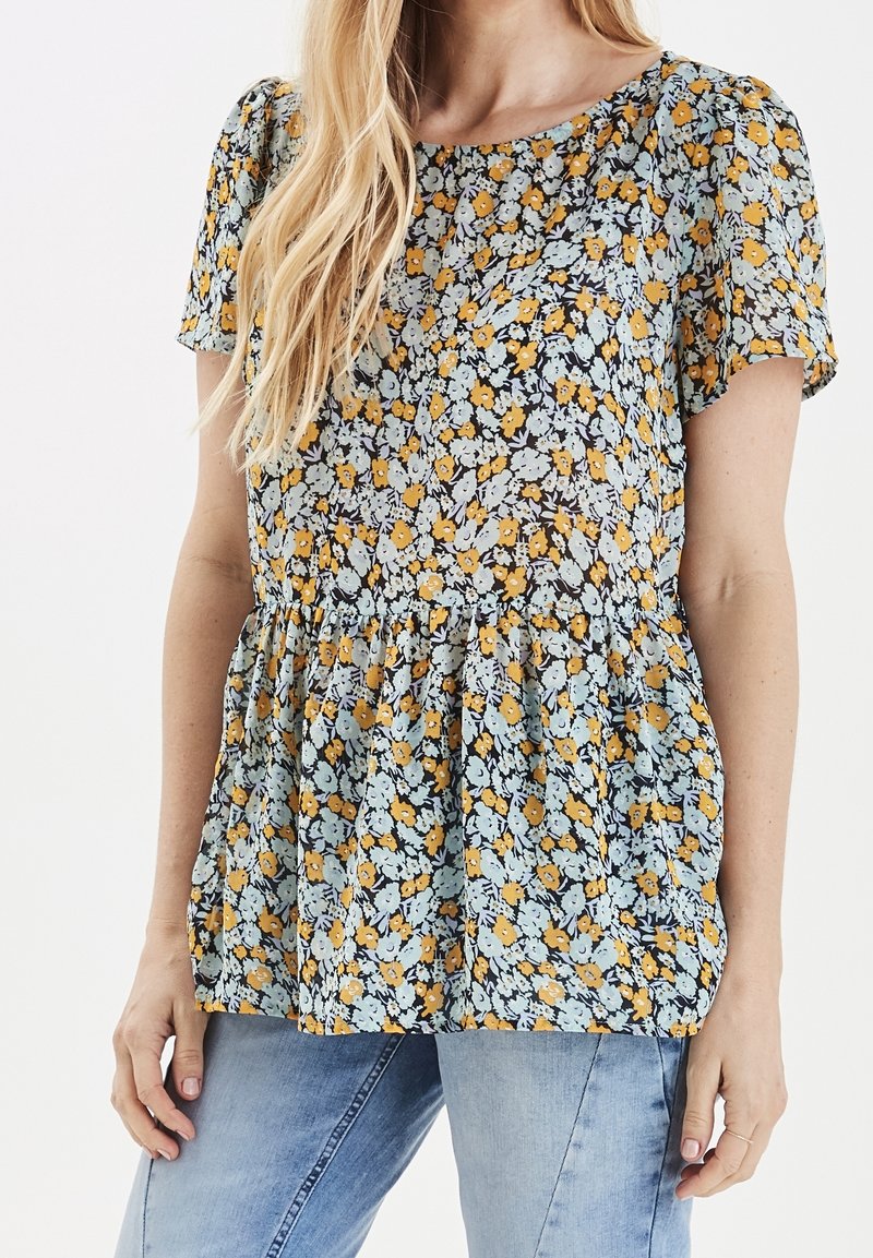 Femme portant un chemisier à manches courtes à fleurs jaunes et bleu clair, associé à un jean bleu clair sur un fond uni.